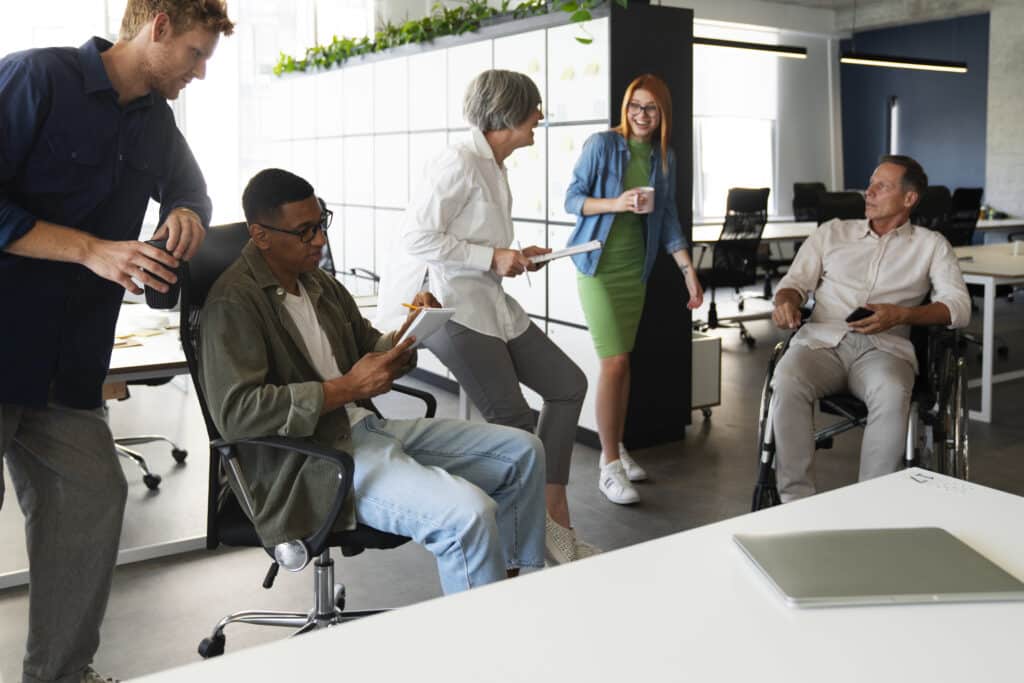 Fünf Personen in einem modernen Büro stehen und sitzen zusammen, unterhalten sich und geben sich gegenseitig Feedback, während sie Notizen und mobile Geräte nutzen.